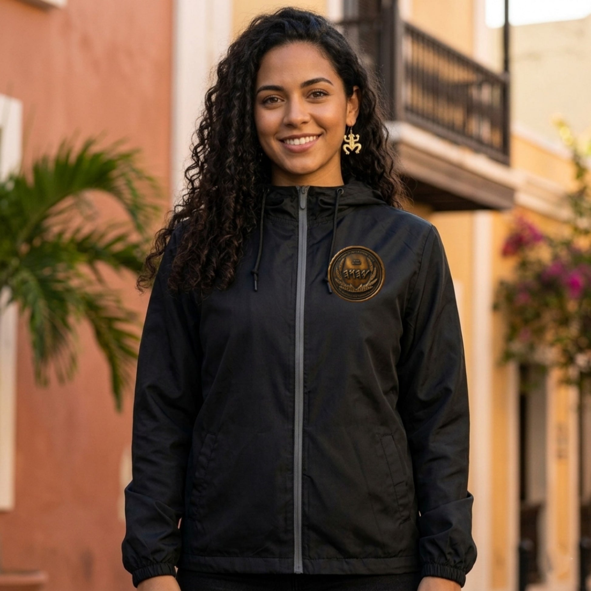 Front view of a woman outdoors wearing a black windbreaker with a small, circular antiqued bronze Paleo-Hebrew script emblem on the left chest.