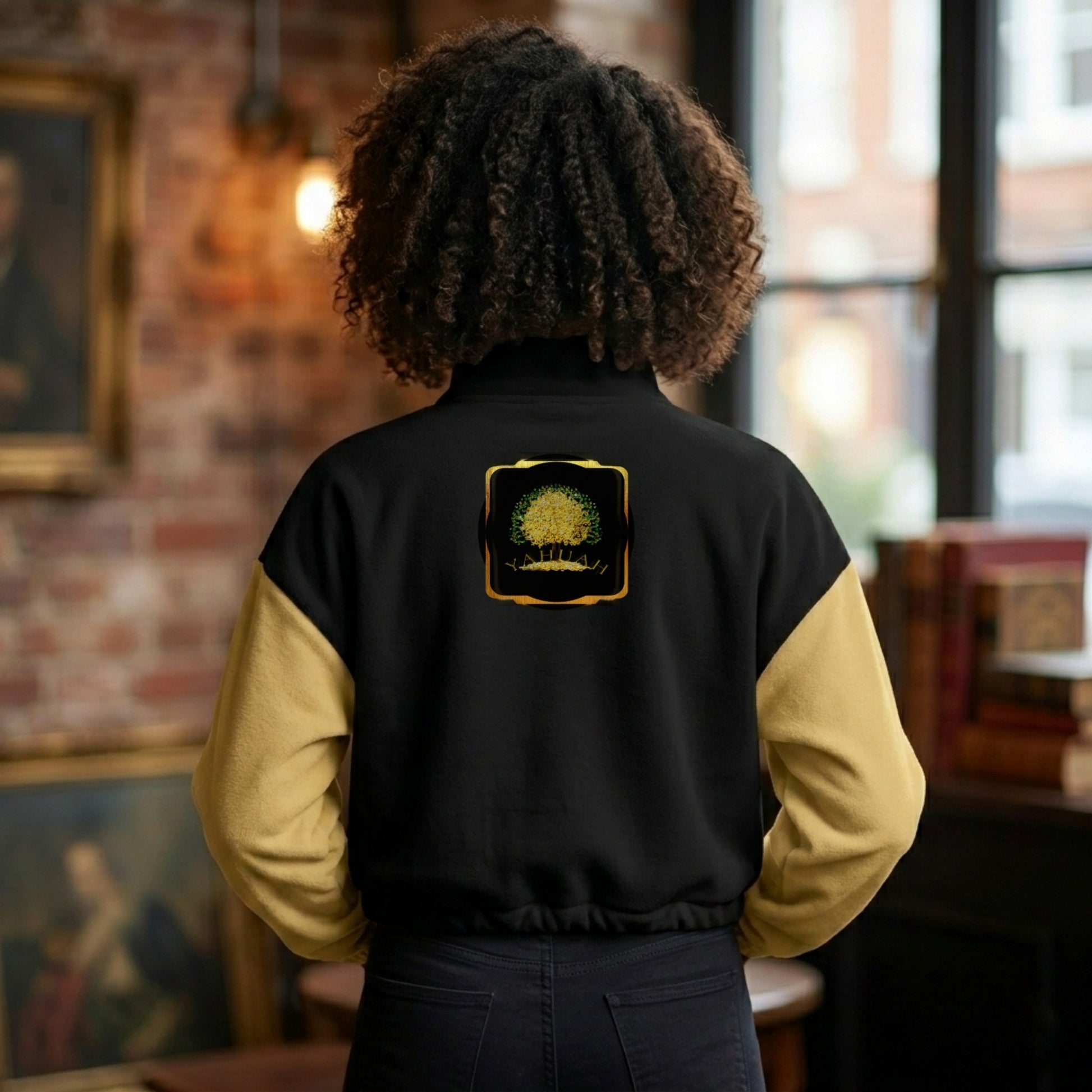 Back view of a woman in a library wearing a black and gold Yahuah: Tree of Life cropped fleece bomber with a golden tree emblem.